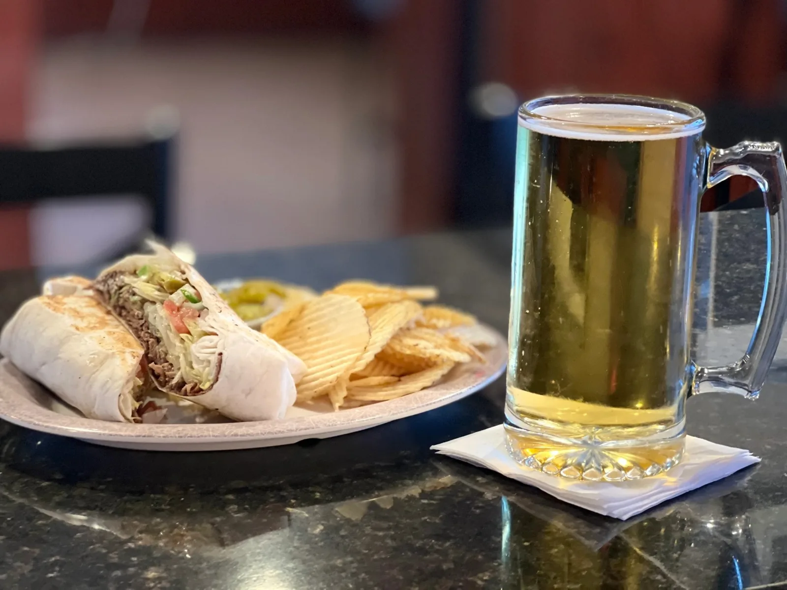 A sandwich with chips on the bar next to a cold mug of draft beer